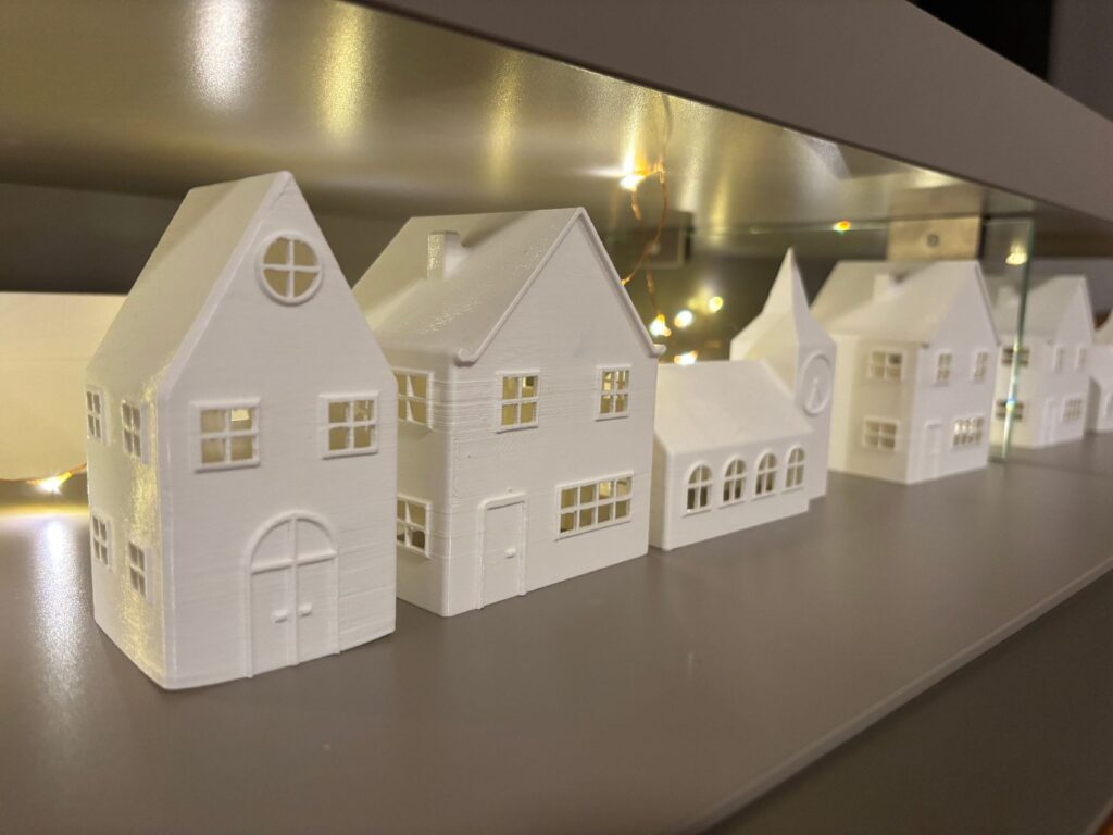 A row of white 3D-printed houses on a shelf, lit from behind by a string of fairy lights