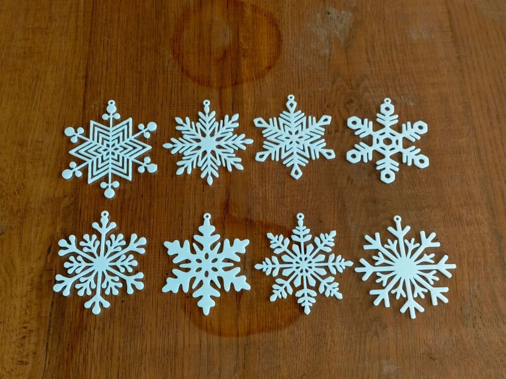 Eight white 3D-printed snowflakes with different patterns, laid out in two rows on a wooden table