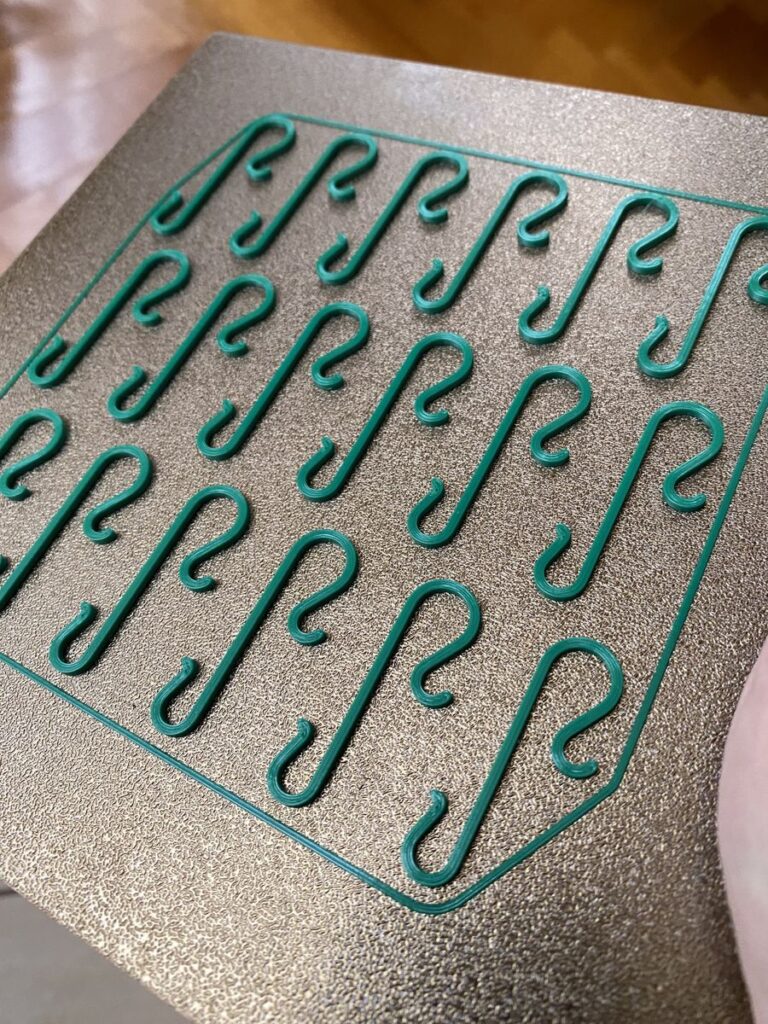 A batch of green 3D-printed Christmas tree hooks on a textured print bed